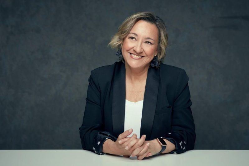 Headshot of Dr. Angela Nieto, in front of a gray background. She's smiling with her fingers interlaced, looking off to the side of the camera, wearing a dark blue blazer and white undershirt.