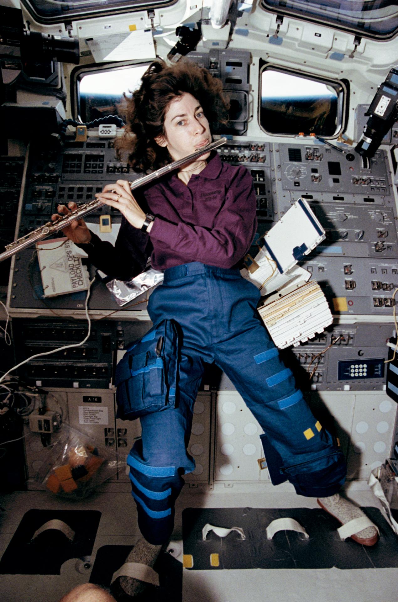 Dr. Ellen Ochoa plays the flute in zero gravity space aboard a NASA spacecraft with books and other objects floating around her on April 17, 1993. Her feet are strapped to the ground.