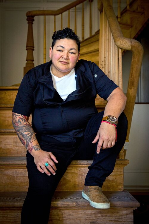 Chef Melissa Araujo poses for a photo on a wooden spiral staircase. She is looking directly at the camera and wearing a dark blue button down shirt over a white undershirt, black pants, a turquoise ring and a watch.