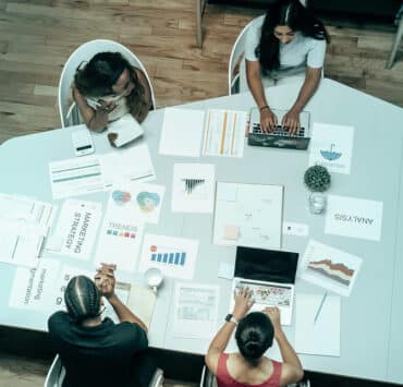 Four women of color sit at a table, working on their computers or on various printed-out finance documents