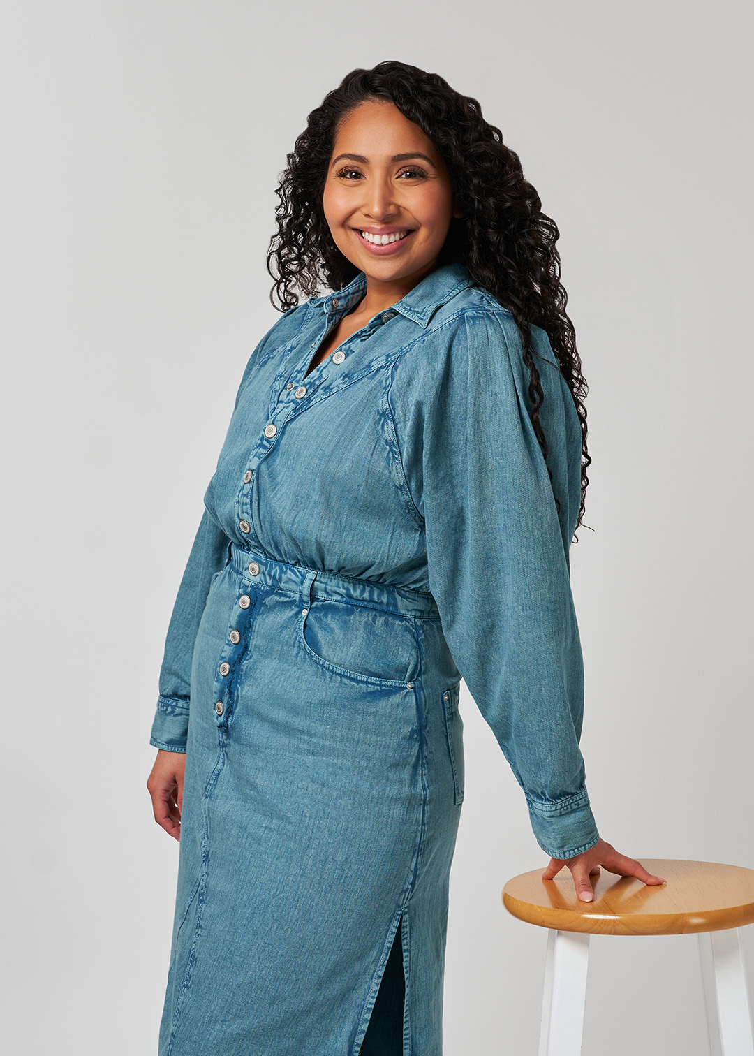 Portrait of American voice actress Kathleen Herles as she wears a denim button-up dress and leans against a stool with a white background