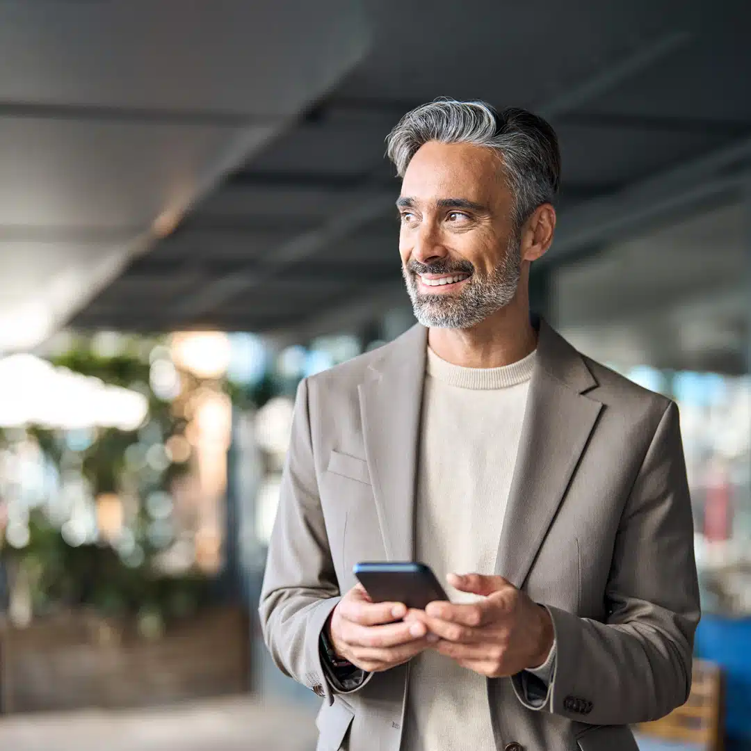 A Latino businessman wearing a taupe blazer smiles and looks off to the side of the camera while holding his iPhone