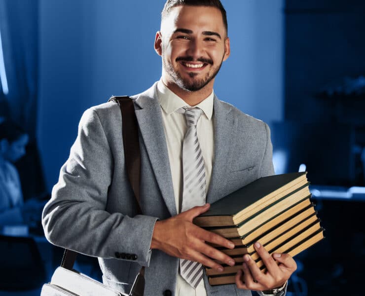 A Latino man holds a stack of books while wearing a gray suit and messenger bag against a blue background