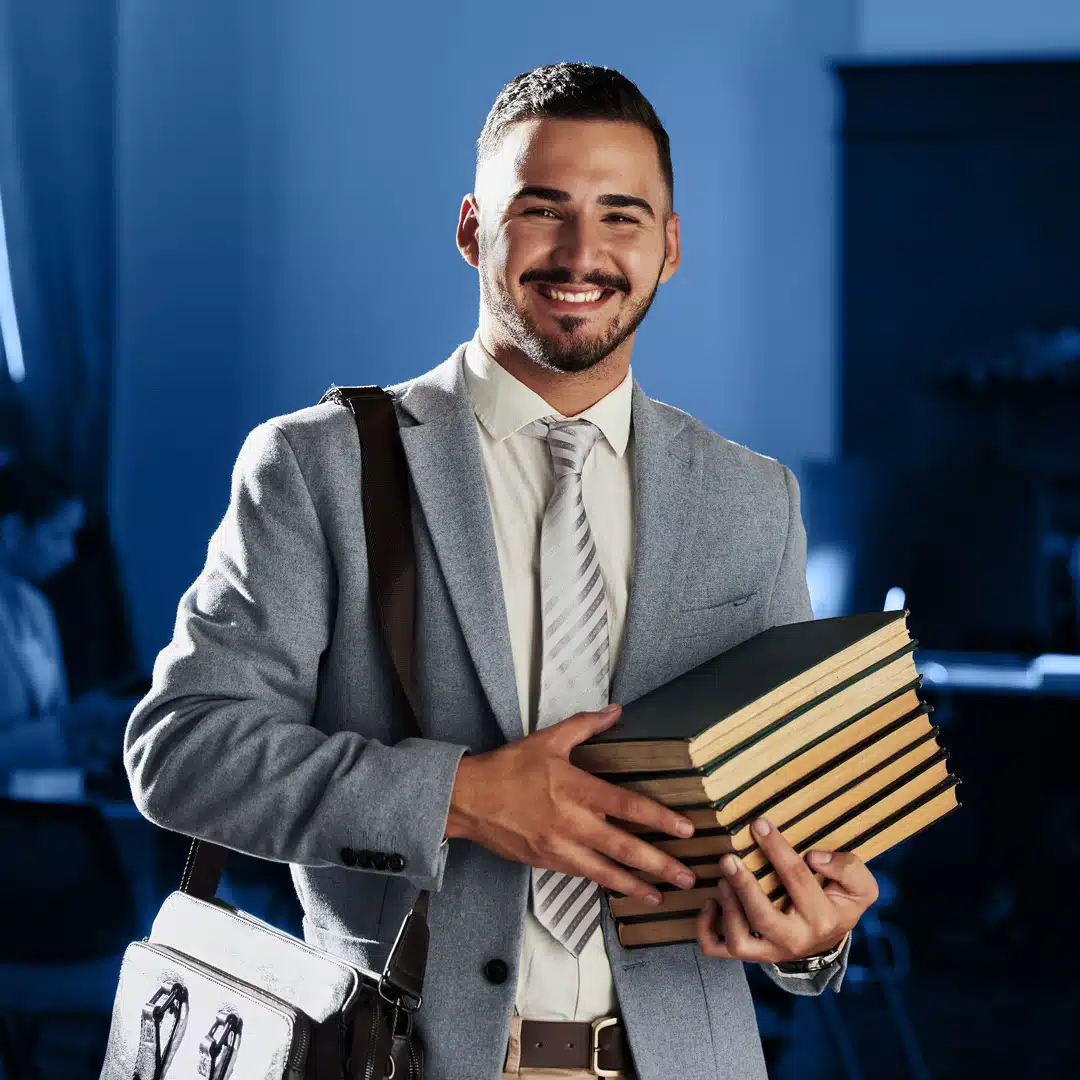 A Latino man holds a stack of books while wearing a gray suit and messenger bag against a blue background