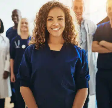 A female healthcare worker smiles, surrounded in the background by other healthcare workers. She is wearing dark blue scrubs.