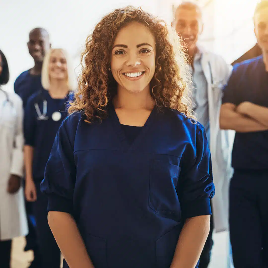 A female healthcare worker smiles, surrounded in the background by other healthcare workers. She is wearing dark blue scrubs.