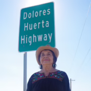 Dolores Huerta wearing a cowboy hat and embroidered blouse, standing beneath a highway sign bearing her name.