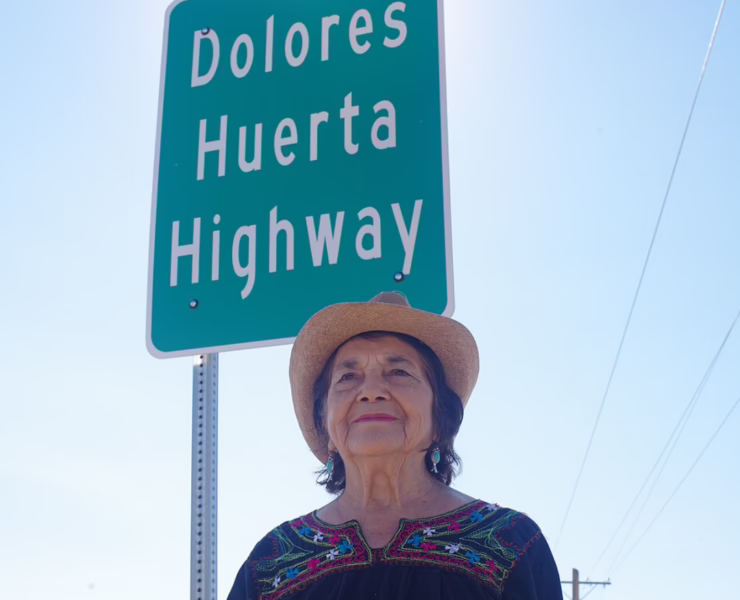 Dolores Huerta wearing a cowboy hat and embroidered blouse, standing beneath a highway sign bearing her name.