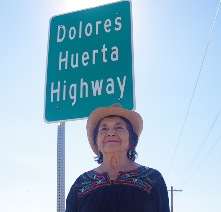 Dolores Huerta wearing a cowboy hat and embroidered blouse, standing beneath a highway sign bearing her name.